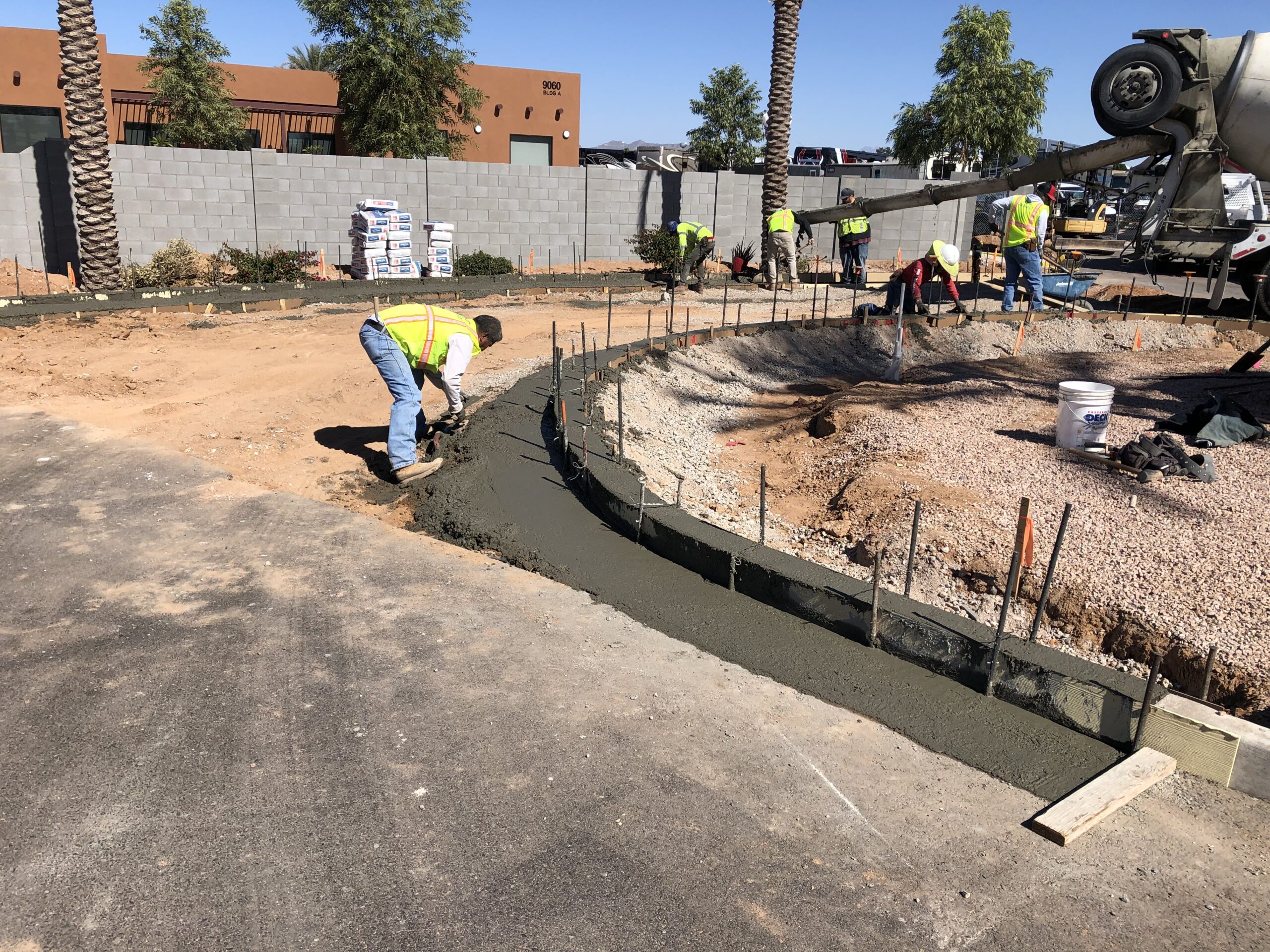 Construction workers in yellow vests pour and smooth wet concrete to form a curved border around a landscaped area, with equipment and materials visible in the background.