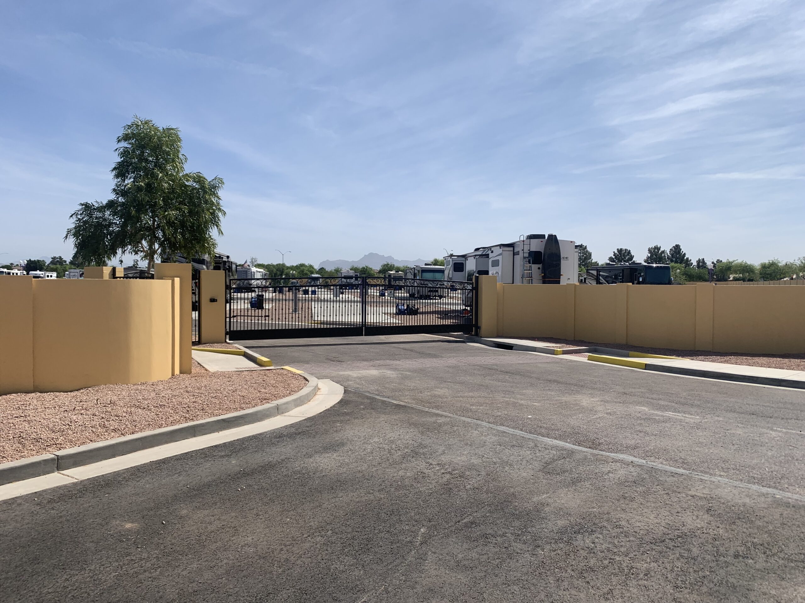 A paved driveway leads to a closed black metal gate with beige walls on each side, enclosing a lot filled with parked RVs under a clear blue sky.