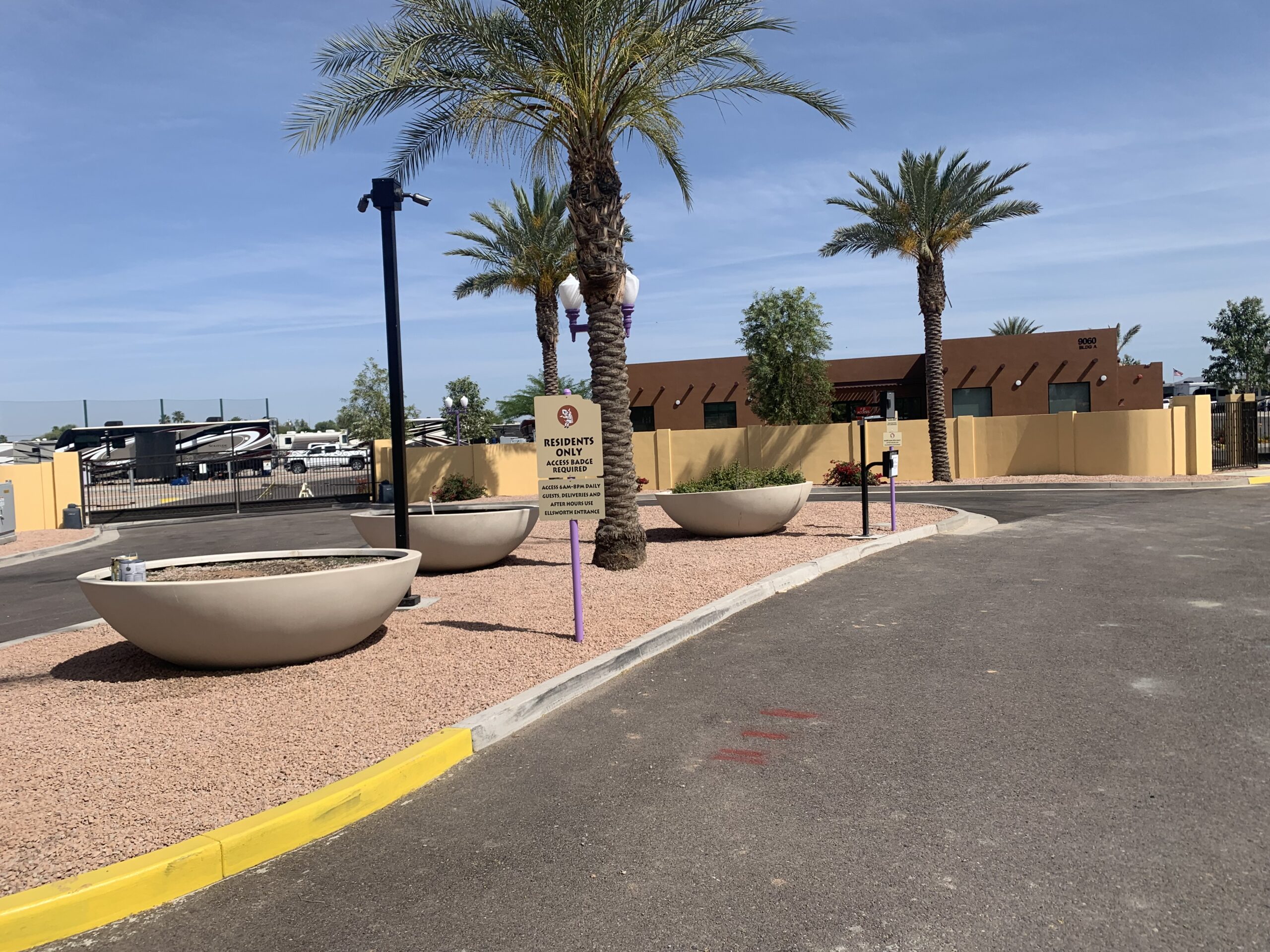 A paved driveway curves past large planters, palm trees, and a sign reading Residents Only in front of a tan building. RVs are visible behind a fence on the left under a clear blue sky.