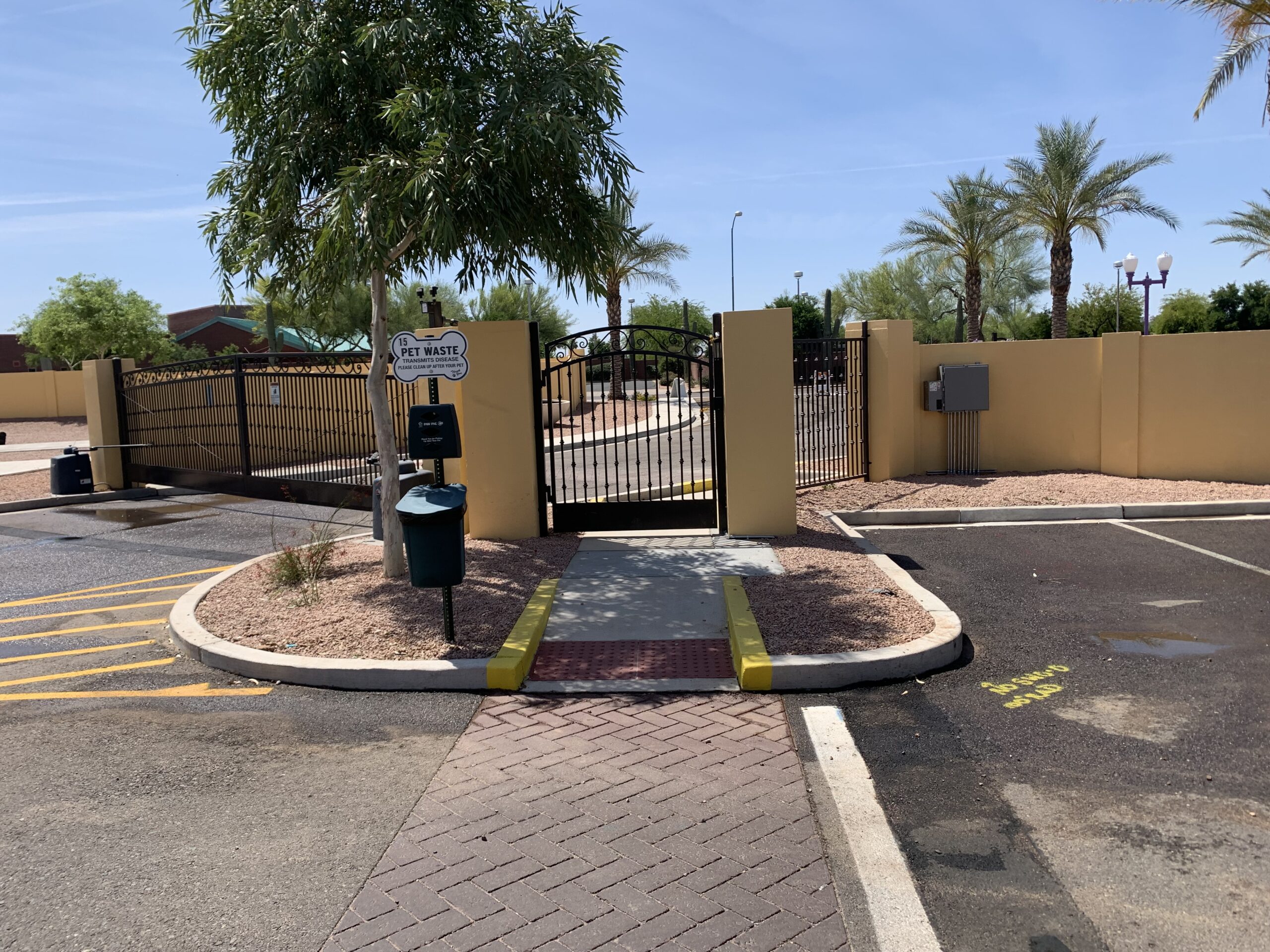 A gated entrance to a pet waste station area with a sign, disposal bin, and a ramp leading up to the gate, surrounded by palm trees and a parking lot under a clear sky.
