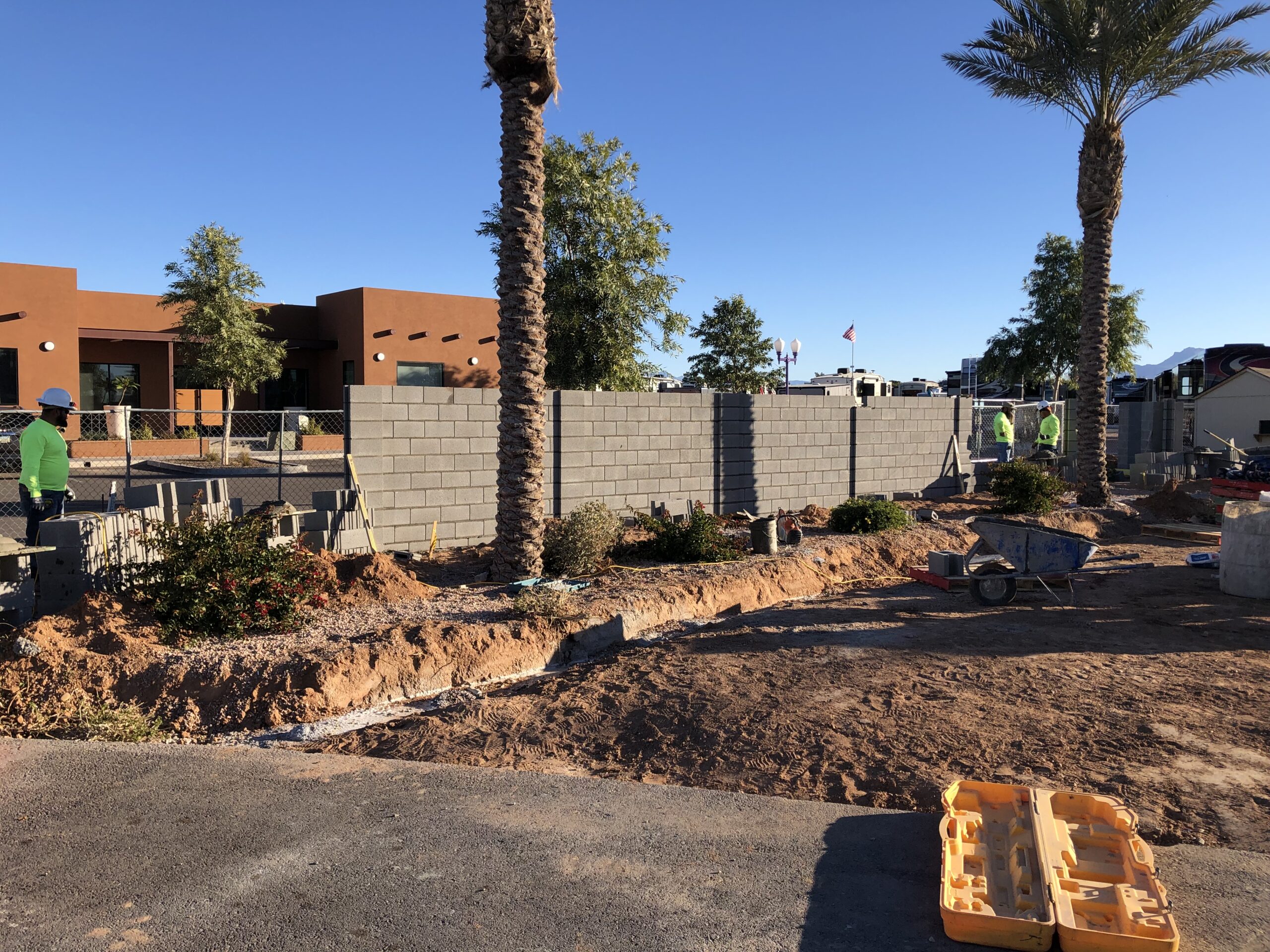 Three workers build a gray cinder block wall behind palm trees and landscaping on a sunny day. Construction tools and equipment are scattered on the ground near the unfinished wall. A modern building is visible in the background.
