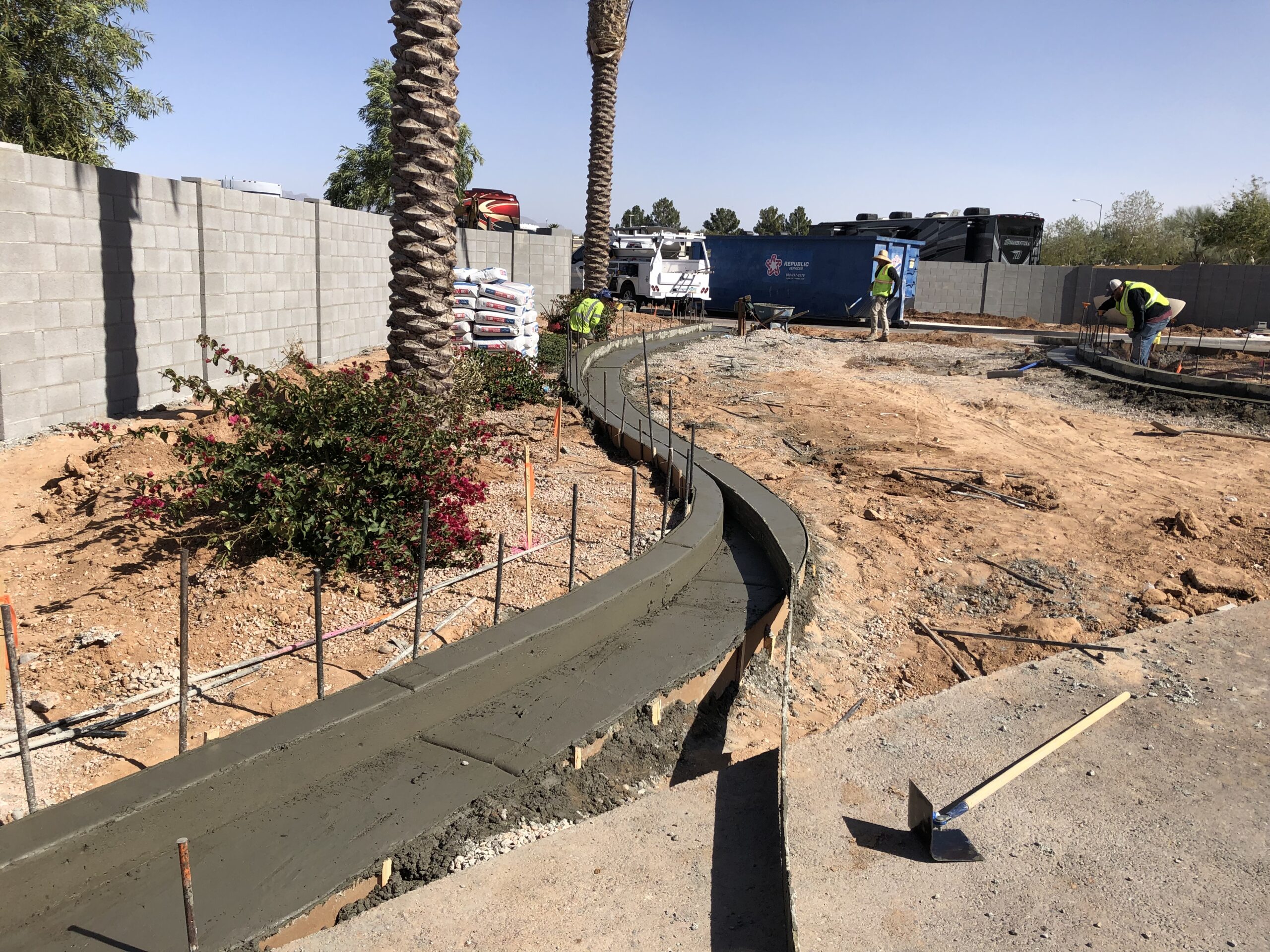 Construction workers pour and shape fresh concrete for a curved pathway in a yard beside a stone wall, with some tools, plants, and palm trees visible under a clear sky.