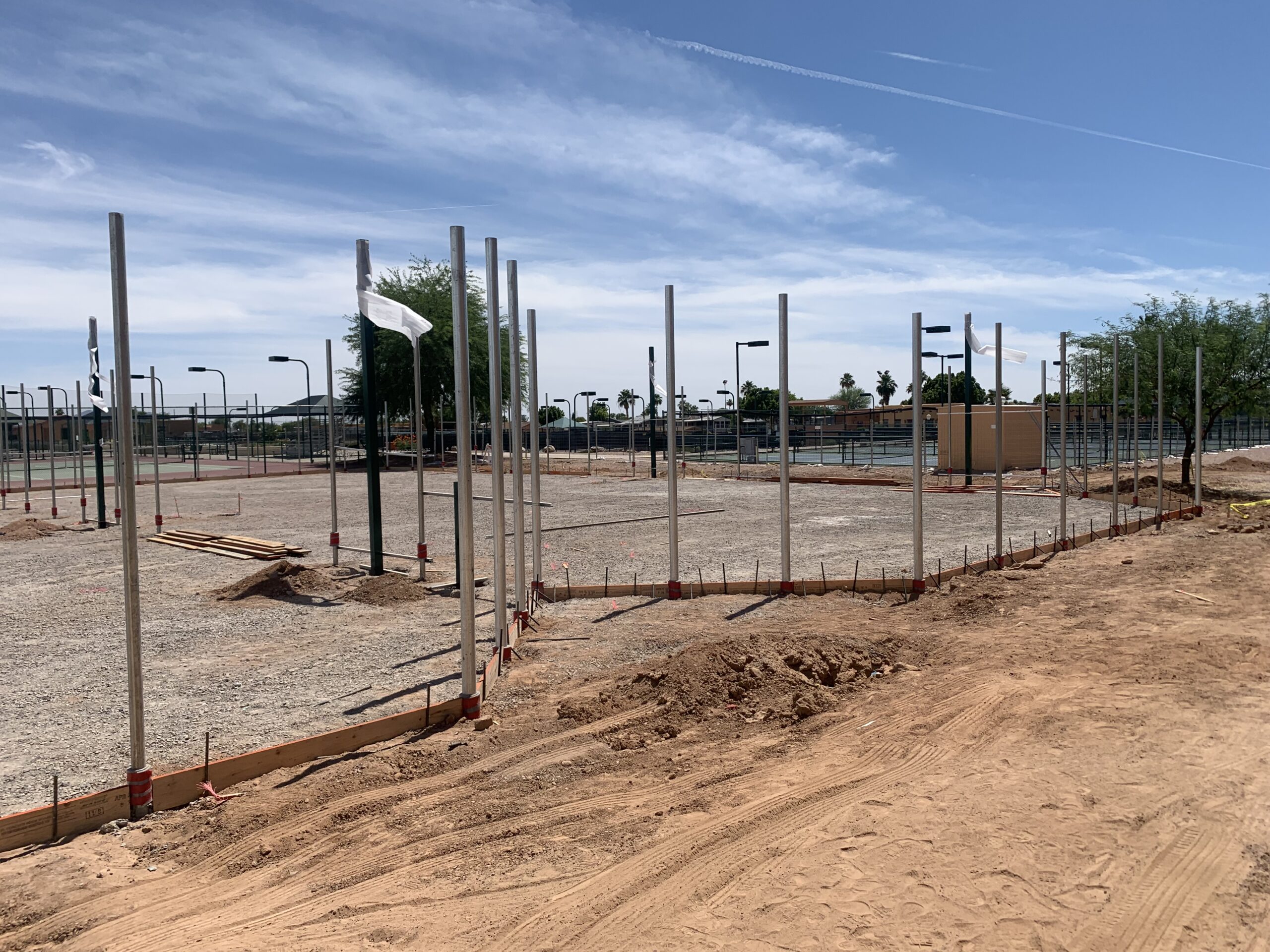 Construction site with metal poles set in the ground, bordered by wooden forms. The area is mostly dirt and gravel, with sports courts and trees visible in the background under a blue sky.