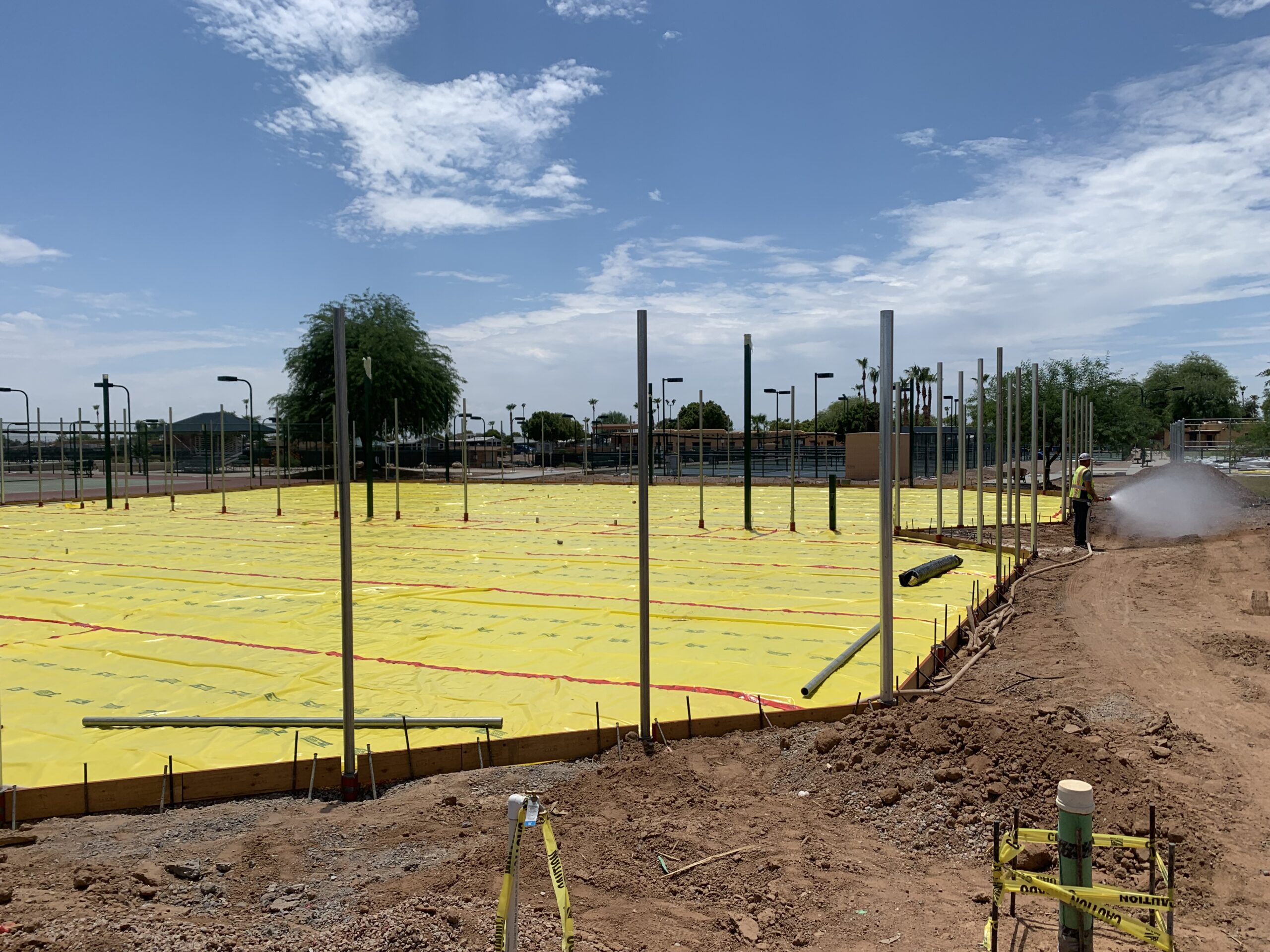 A construction site with a foundation covered in yellow plastic sheeting, several upright metal poles, exposed soil, and caution tape; a worker sprays water at the edge under a partly cloudy sky.