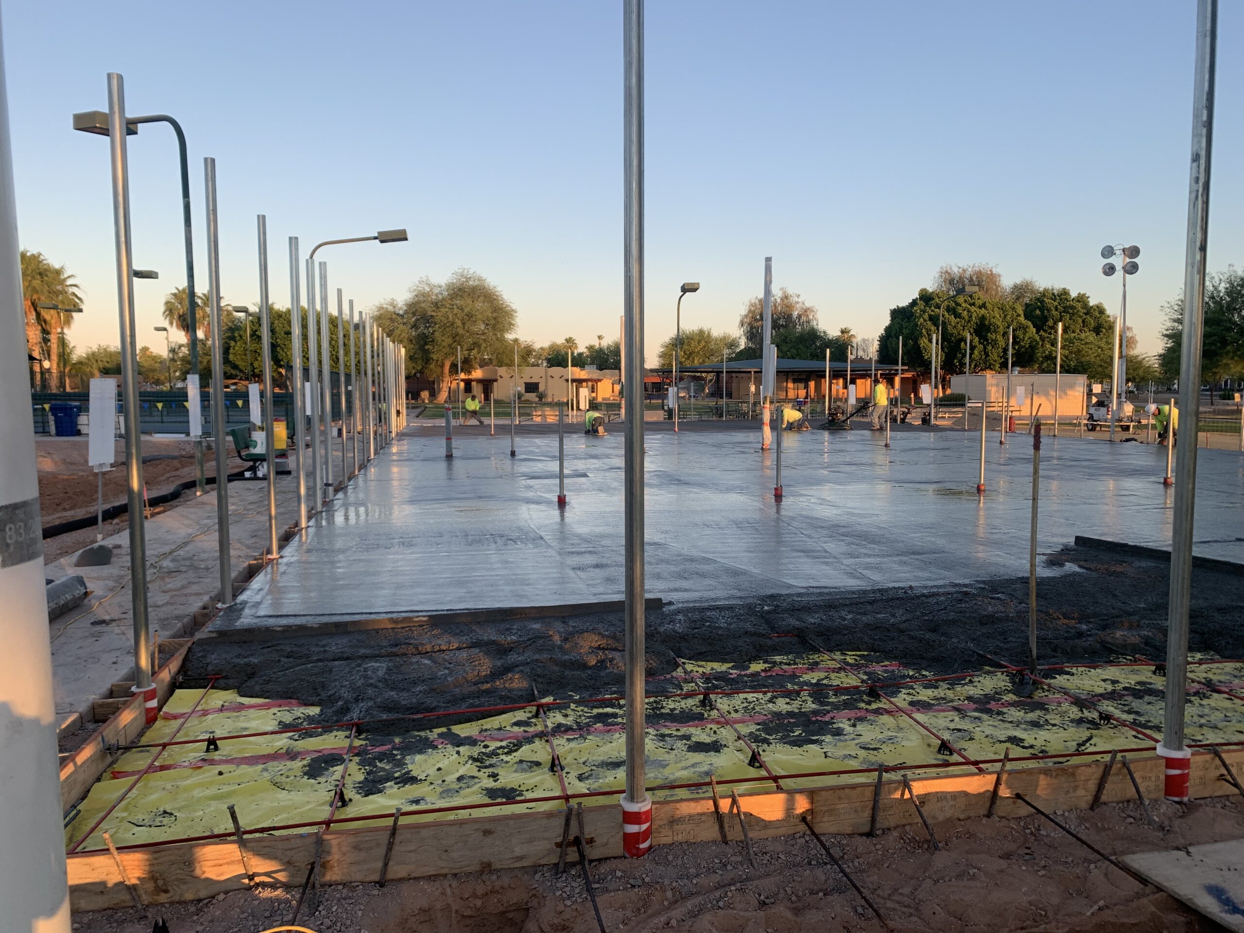 Construction site at sunset showing a large, freshly poured concrete slab foundation with metal poles around it. Workers and equipment are visible in the background, and trees line the site perimeter.