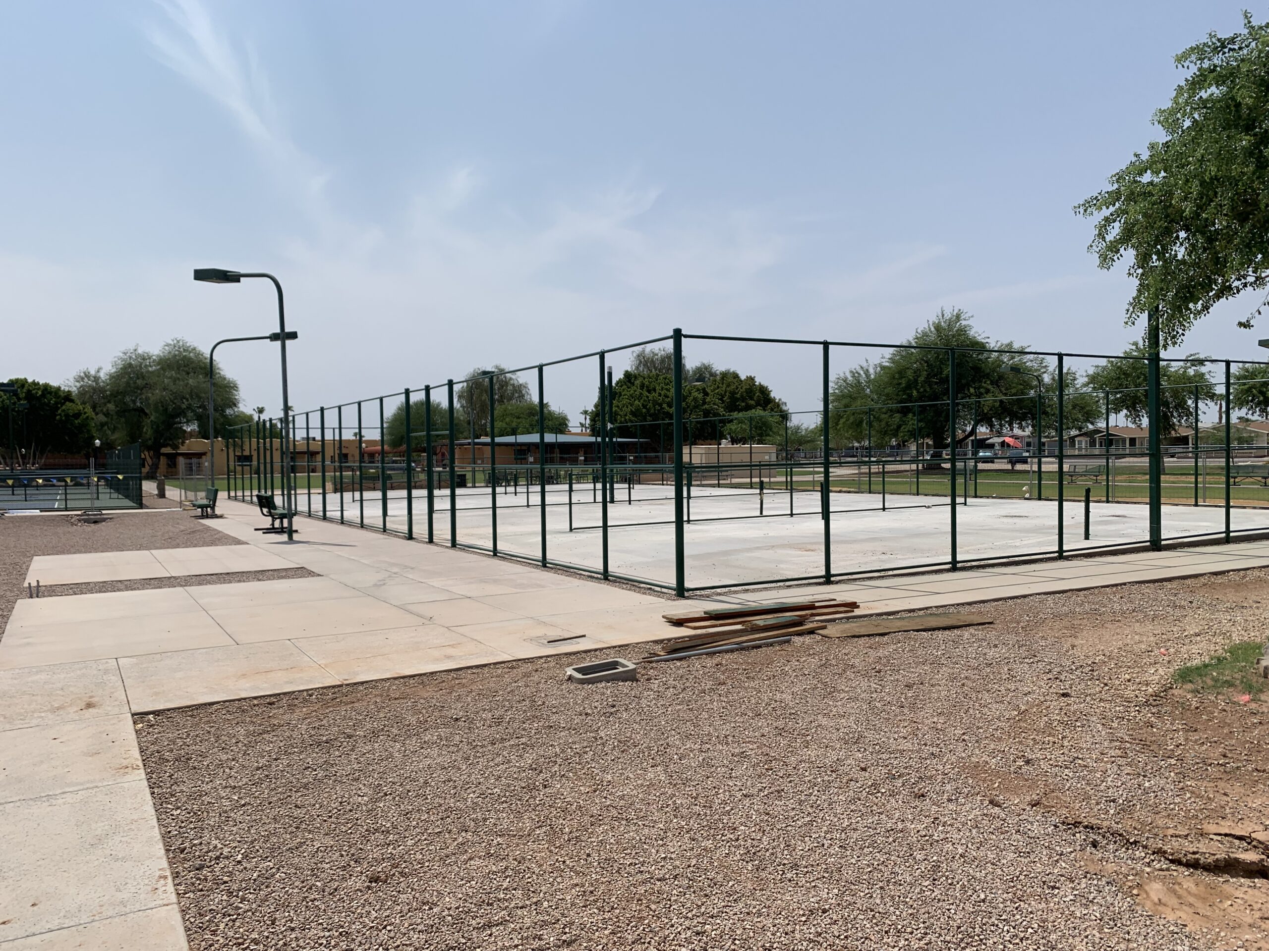 Outdoor tennis and pickleball courts surrounded by a green metal fence in a park, with benches, trees, and scattered construction materials nearby on a sunny day.