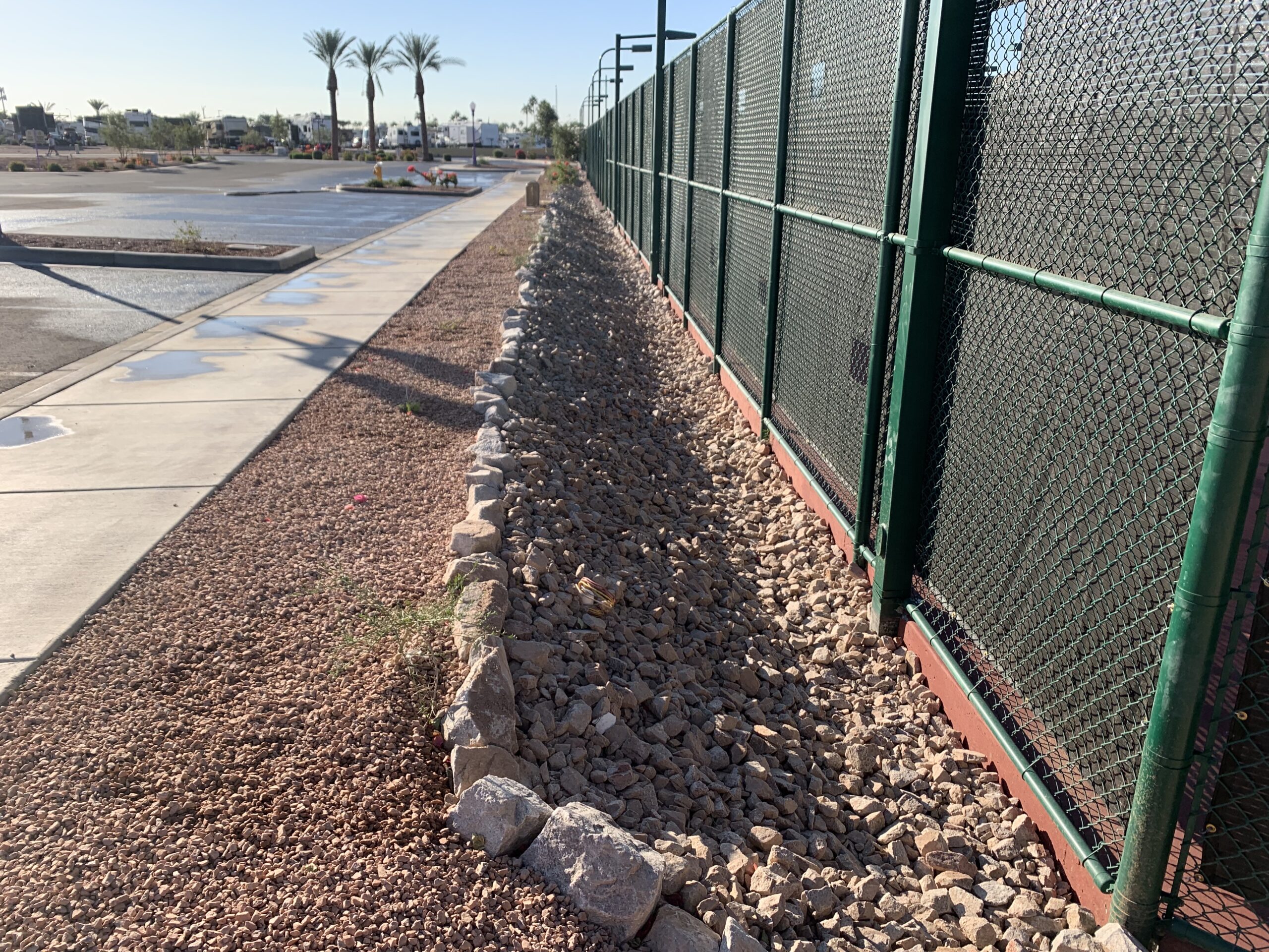 A green chain-link fence runs along a gravel-filled ditch bordered by rocks, with a sidewalk and empty parking lot to the left. Palm trees and distant buildings are visible under a clear blue sky.