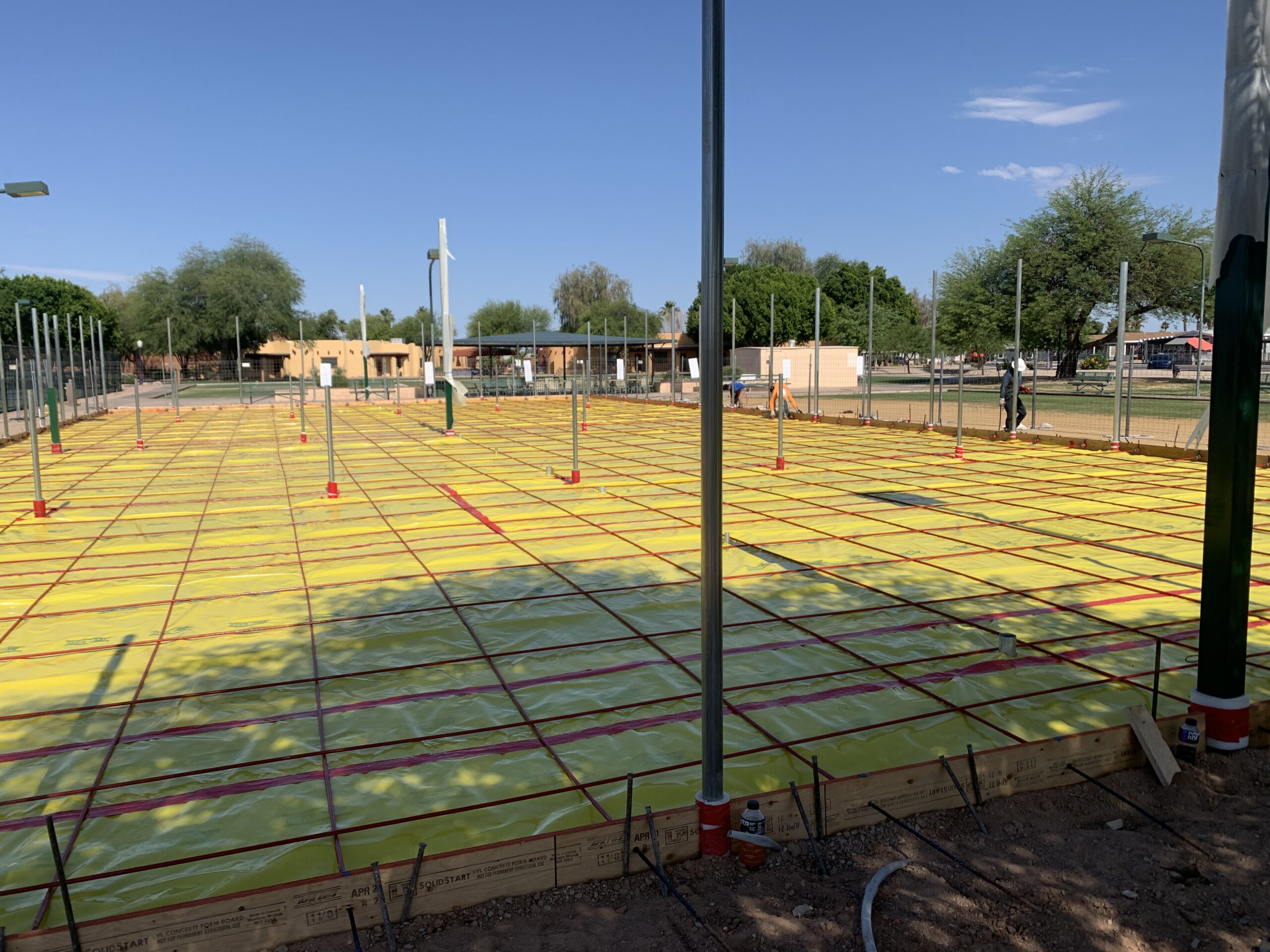 A construction site with a flat area covered in yellow plastic sheeting and red grid lines, surrounded by metal poles. Trees and buildings are visible in the background under a clear blue sky.