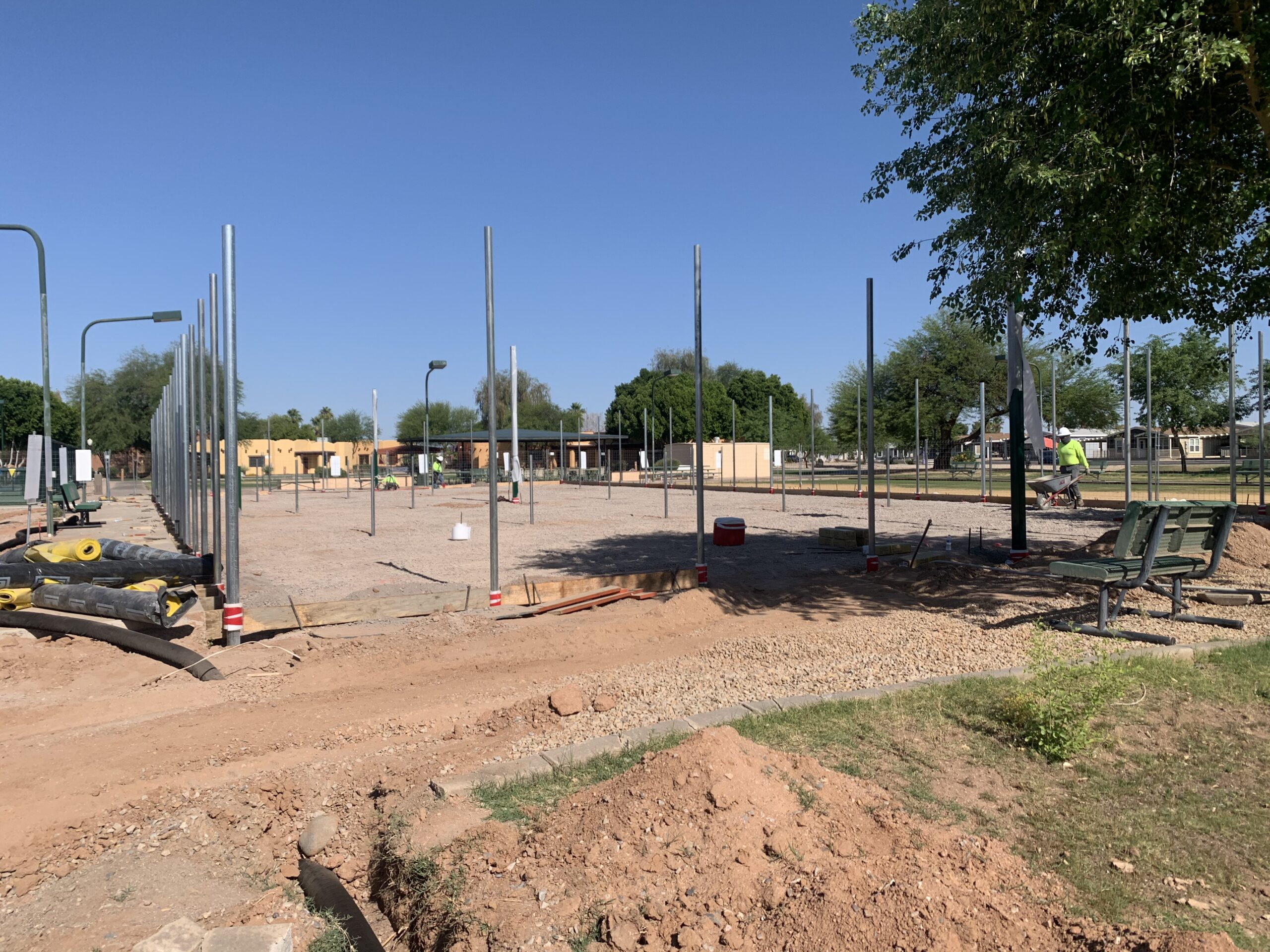 A construction site with metal poles set in the ground, surrounded by dirt and gravel. A worker in a safety vest stands nearby, and some benches and trees are visible in the background under a clear blue sky.