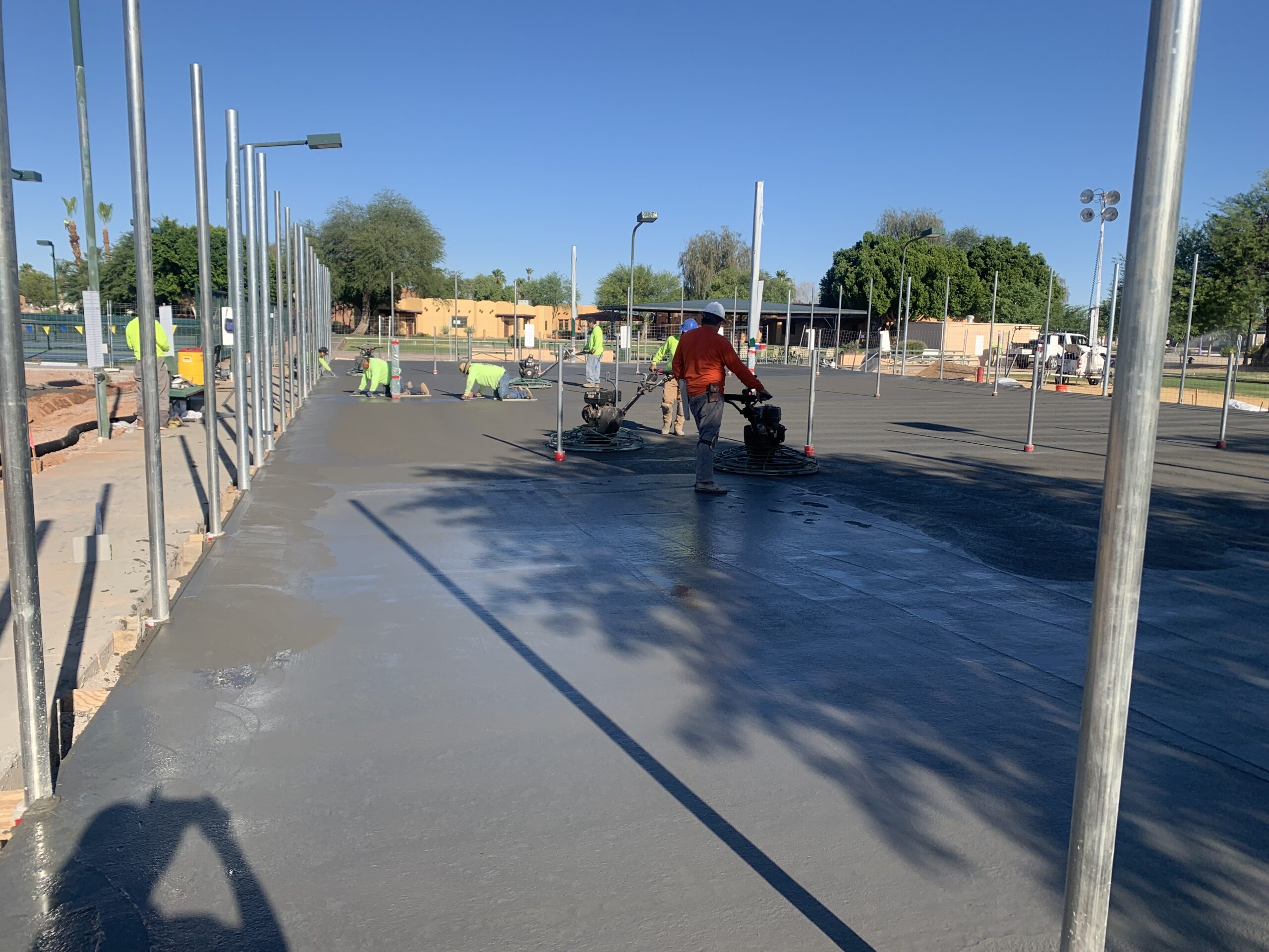Workers smooth freshly poured concrete on a large outdoor sports court under a clear blue sky, surrounded by metal poles and construction equipment. Shadows and trees are visible in the background.