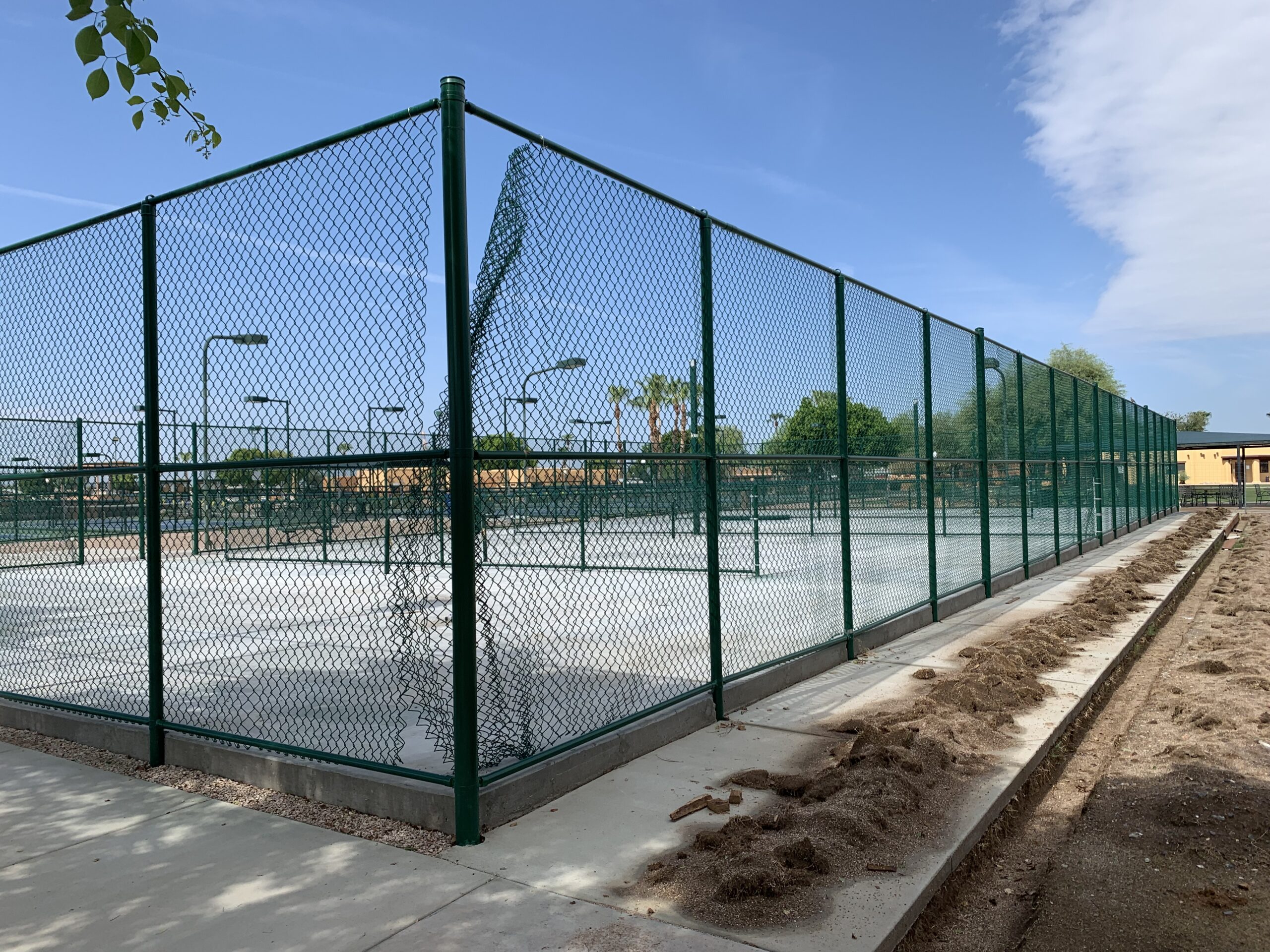 A fenced outdoor tennis court with a section of damaged chain-link fence. The court appears empty, and there is a dirt trench alongside the sidewalk bordering the fence. Trees and blue sky are visible in the background.