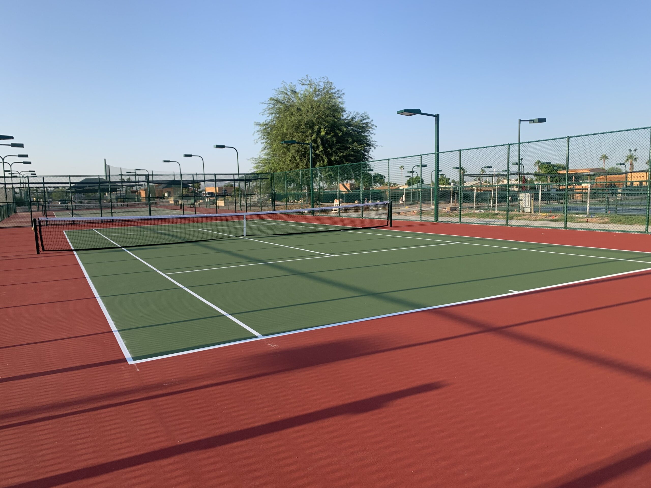 An empty outdoor pickleball court with red and green surfaces, surrounded by a tall chain-link fence and light poles, under a clear blue sky with a few trees and buildings in the background.