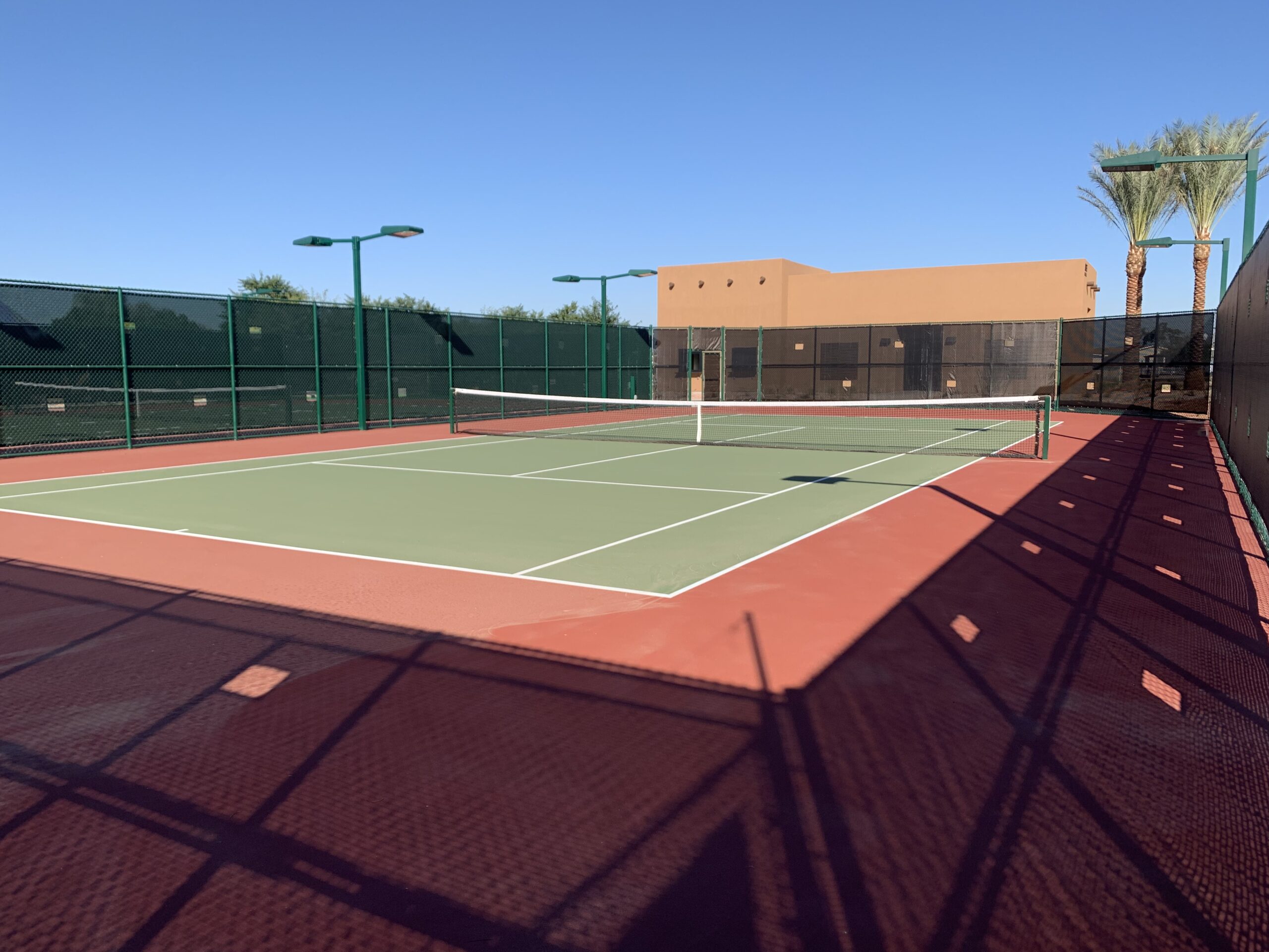 A tennis court with a green and red surface is empty under a clear blue sky, surrounded by a black fence. In the background, there’s a tan building and several palm trees.