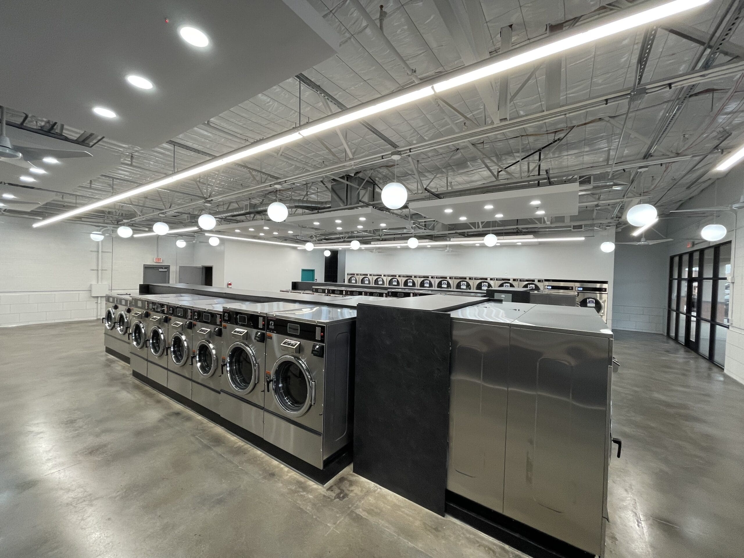 A modern laundromat offering laundry services with rows of stainless steel washing machines and dryers on a polished concrete floor, under bright ceiling lights and exposed piping.