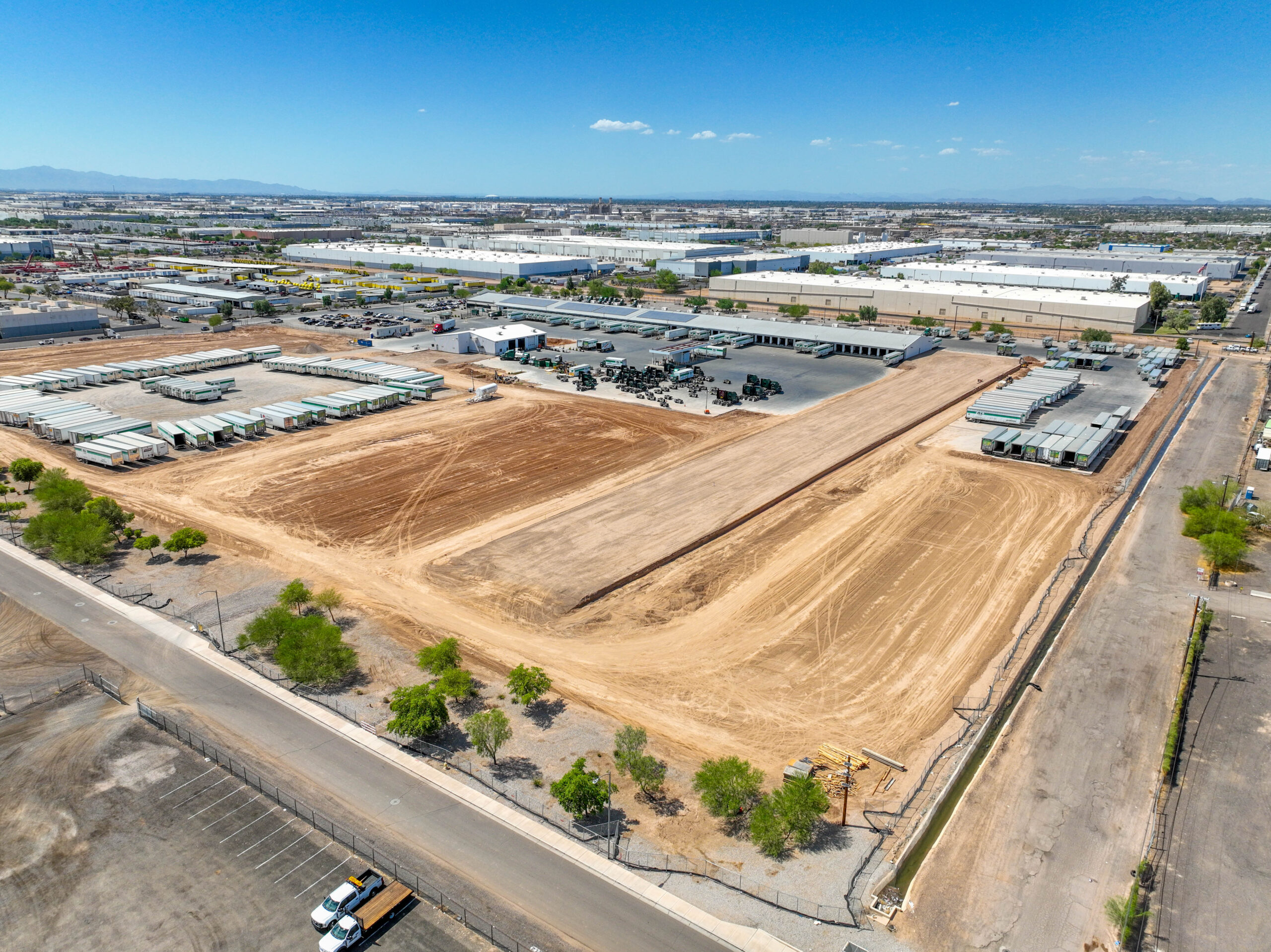 A large construction site with leveled dirt, surrounded by roads and trees, sits next to industrial buildings and parked trucks from R+L Carriers under a clear blue sky—an active hub of logistics and freight shipping.