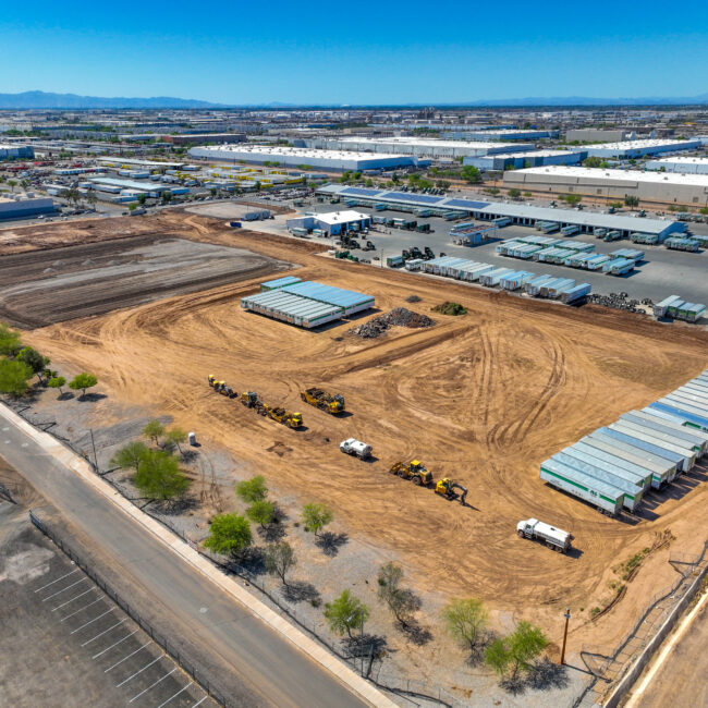 Aerial view of a large industrial construction site with earthmoving equipment, storage containers, and warehouses—ideal for logistics operations. The ground is mostly dirt, with scattered vehicles and nearby industrial buildings under a clear blue sky.