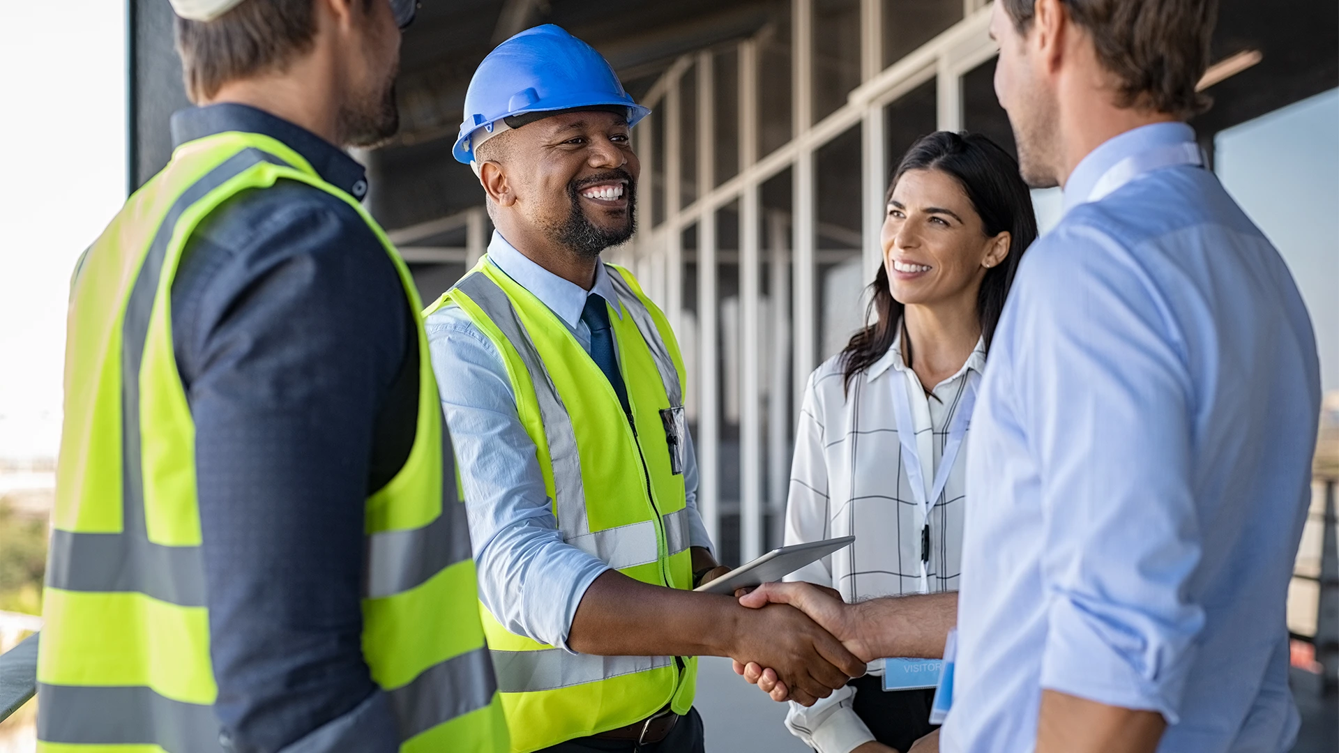 A smiling construction worker in a safety vest and blue helmet shakes hands with a person in business attire while two colleagues, a man in a vest and a woman holding a tablet, stand nearby and watch.
