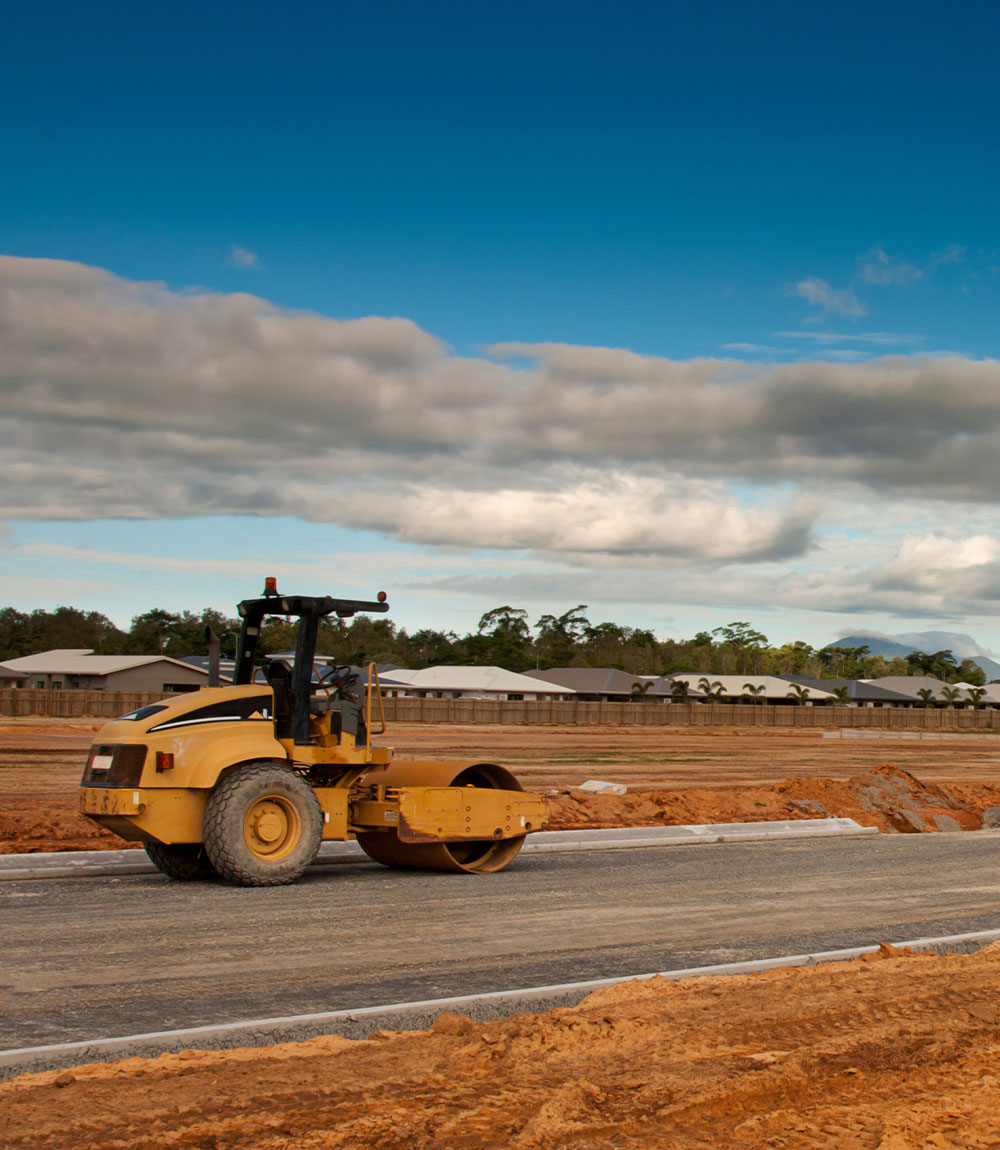 A yellow road roller flattens a new road surface at a construction site under a blue sky with clouds, with houses and trees visible in the background.