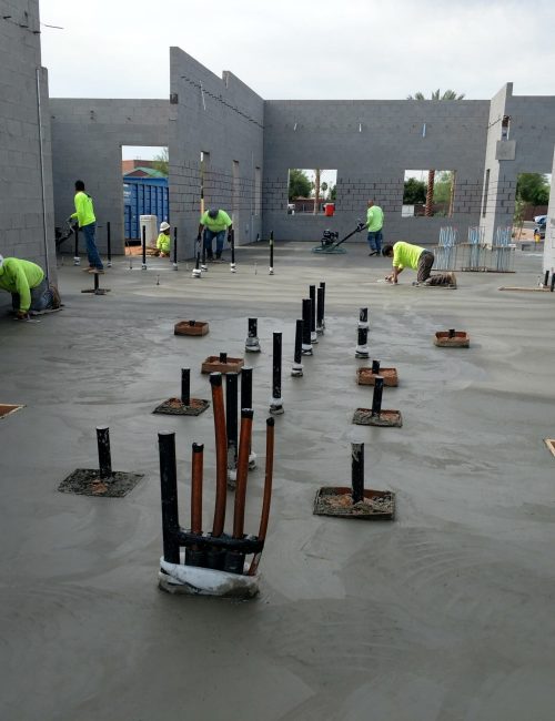 Construction workers in green shirts smooth wet concrete on a building floor with unfinished gray cinder block walls; exposed pipes and plumbing fixtures protrude from the floor.